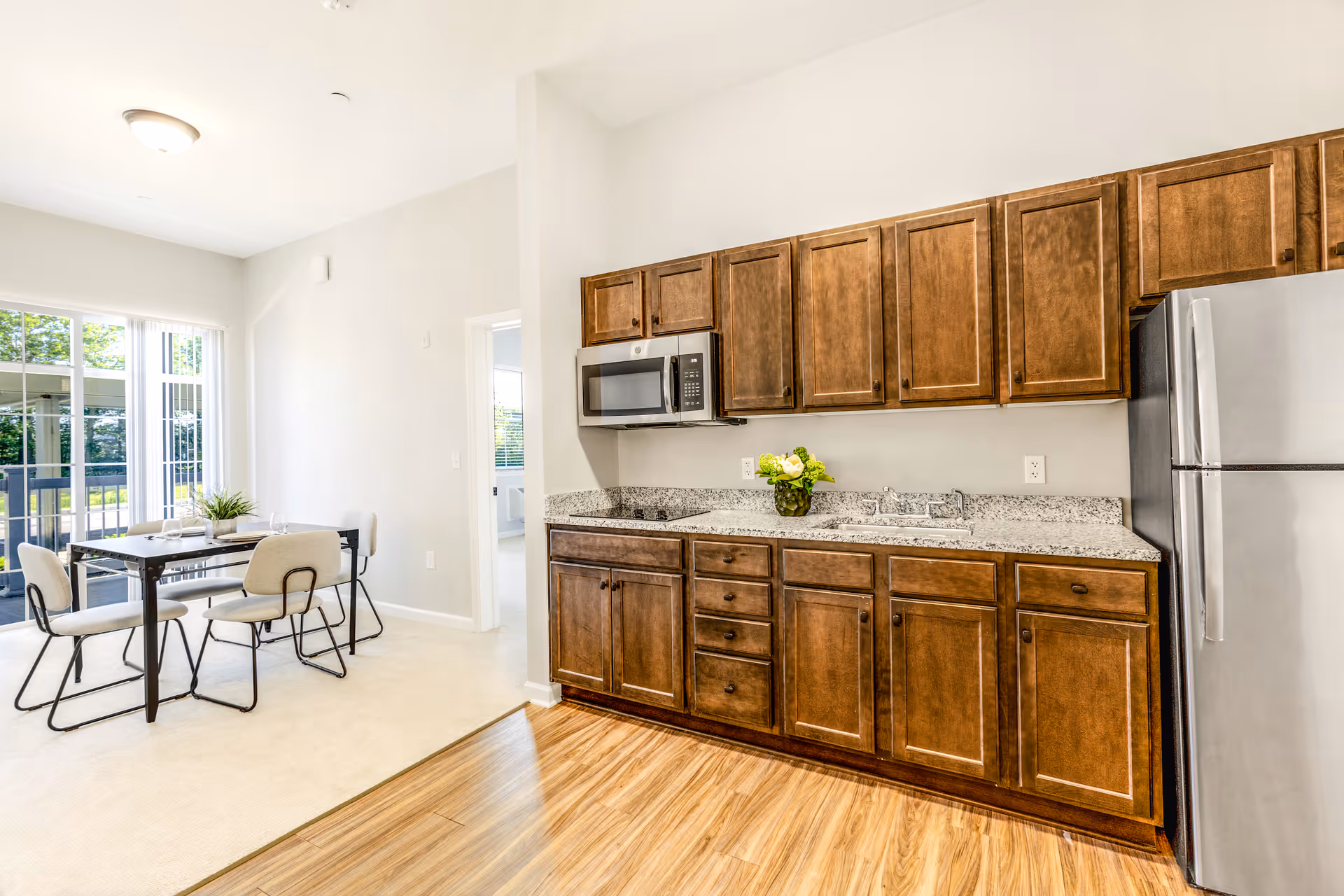 Bright kitchen area with wooden cabinets, granite countertops, a stainless steel microwave, and refrigerator. Adjacent to the kitchen is a dining area with a table and four chairs near large windows letting in natural light.