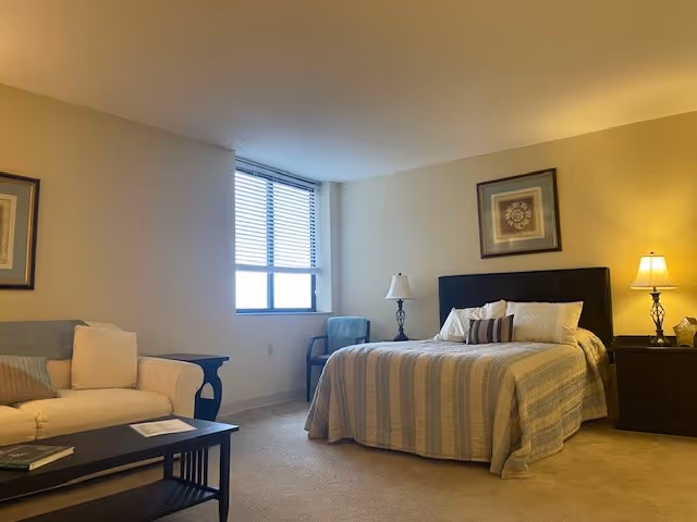A well-lit bedroom in a senior living facility featuring a large bed with striped bedding and multiple pillows, a nightstand with a lamp, a framed picture above the bed, a window with blinds, a cushioned chair near the window, a beige sofa with pillows, and a coffee table with books and papers.
