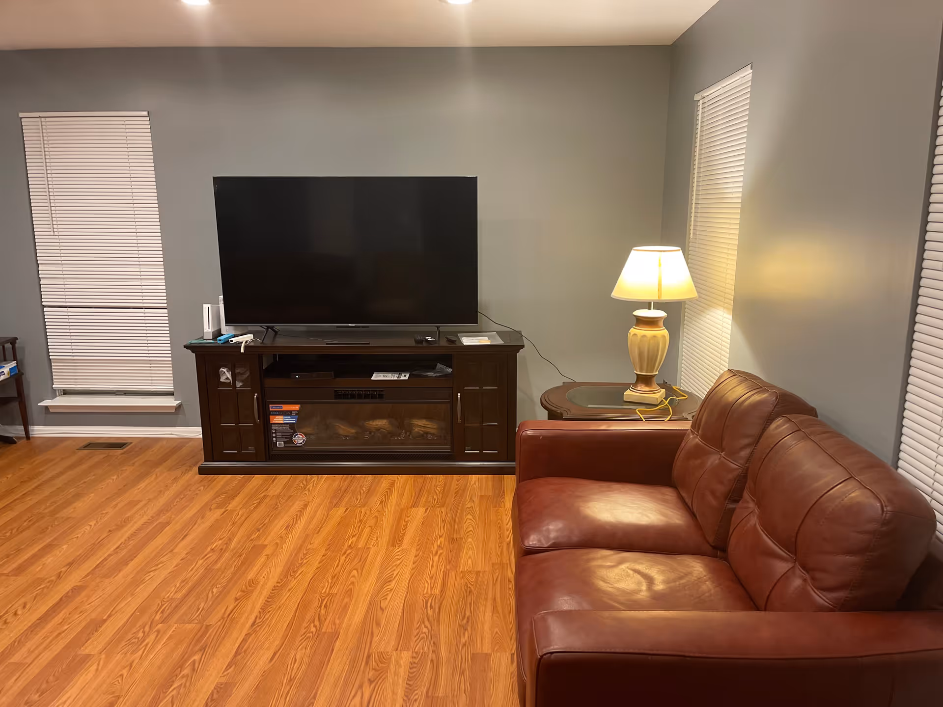 A living room with a brown leather couch on the right, a wooden side table with a lamp, and a large flat-screen TV on a dark wooden TV stand with an electric fireplace. The room has light gray walls, two windows with closed blinds, and wood laminate flooring.