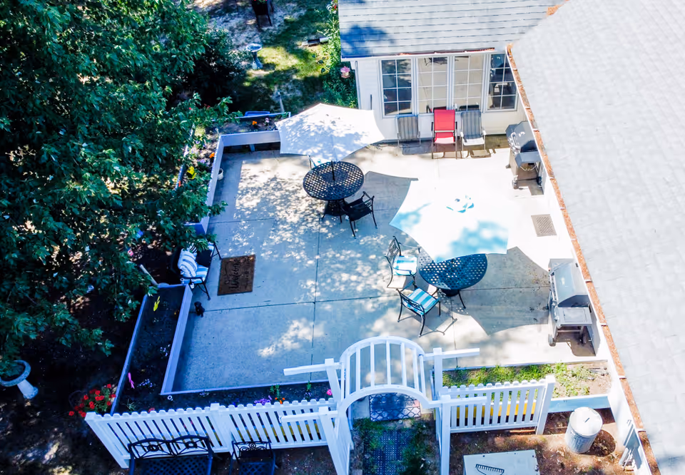 Aerial view of a fenced outdoor patio with tables, umbrellas, chairs, and grills next to a building.