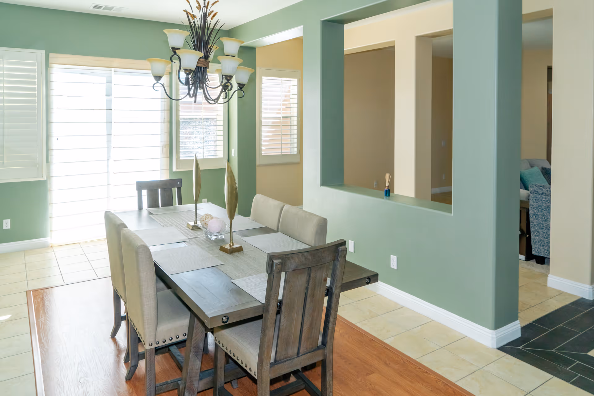 A bright dining room with a wooden dining table set for six, featuring beige cushioned chairs and a decorative centerpiece. The room has green walls with a large window covered by white blinds, a chandelier hanging above the table, and a pass-through window opening to an adjacent room.