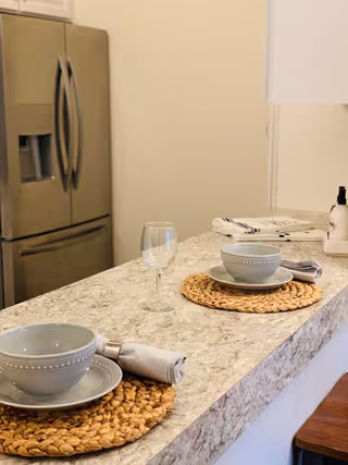 A kitchen counter set for two with woven placemats, gray bowls and plates, cloth napkins, and a wine glass. In the background, there is a stainless steel refrigerator and some folded kitchen towels on the counter.