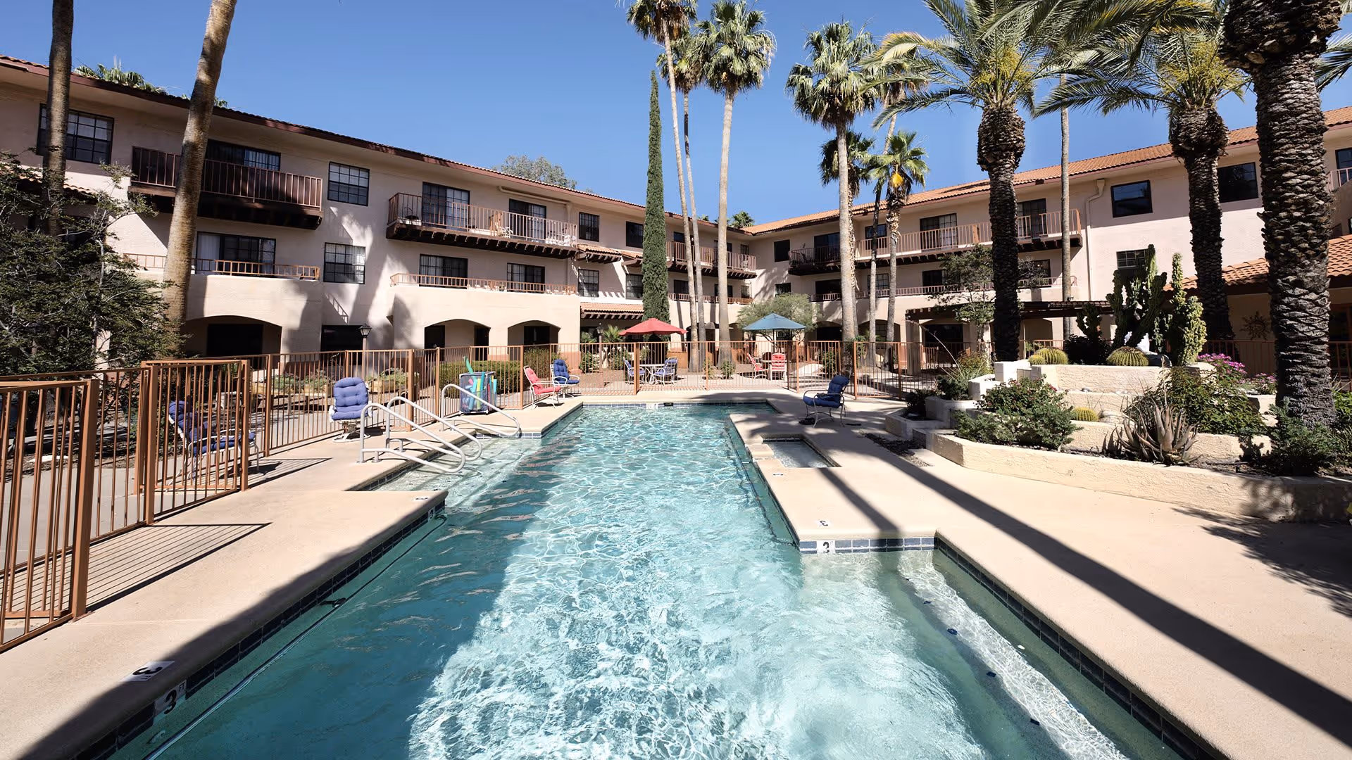Outdoor swimming pool area surrounded by a three-story building with balconies, palm trees, lounge chairs, and umbrellas under a clear blue sky.