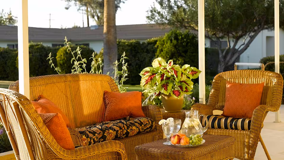 Outdoor patio area with wicker furniture including a loveseat and two armchairs with orange cushions, a wicker coffee table with a pitcher of water, glasses, and a plate of fruit, and a potted plant in the background.
