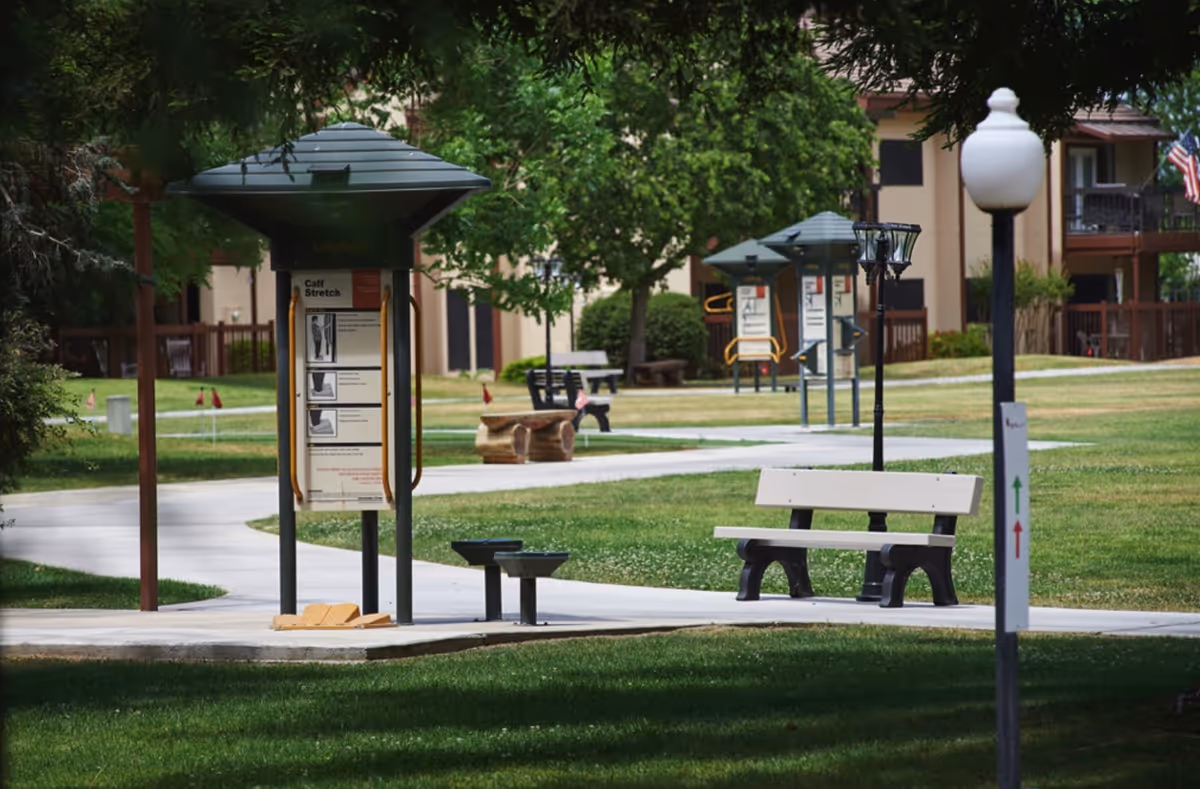 Outdoor exercise area in a senior living facility with multiple exercise stations, benches, paved walkways, green grass, trees, and residential buildings in the background.