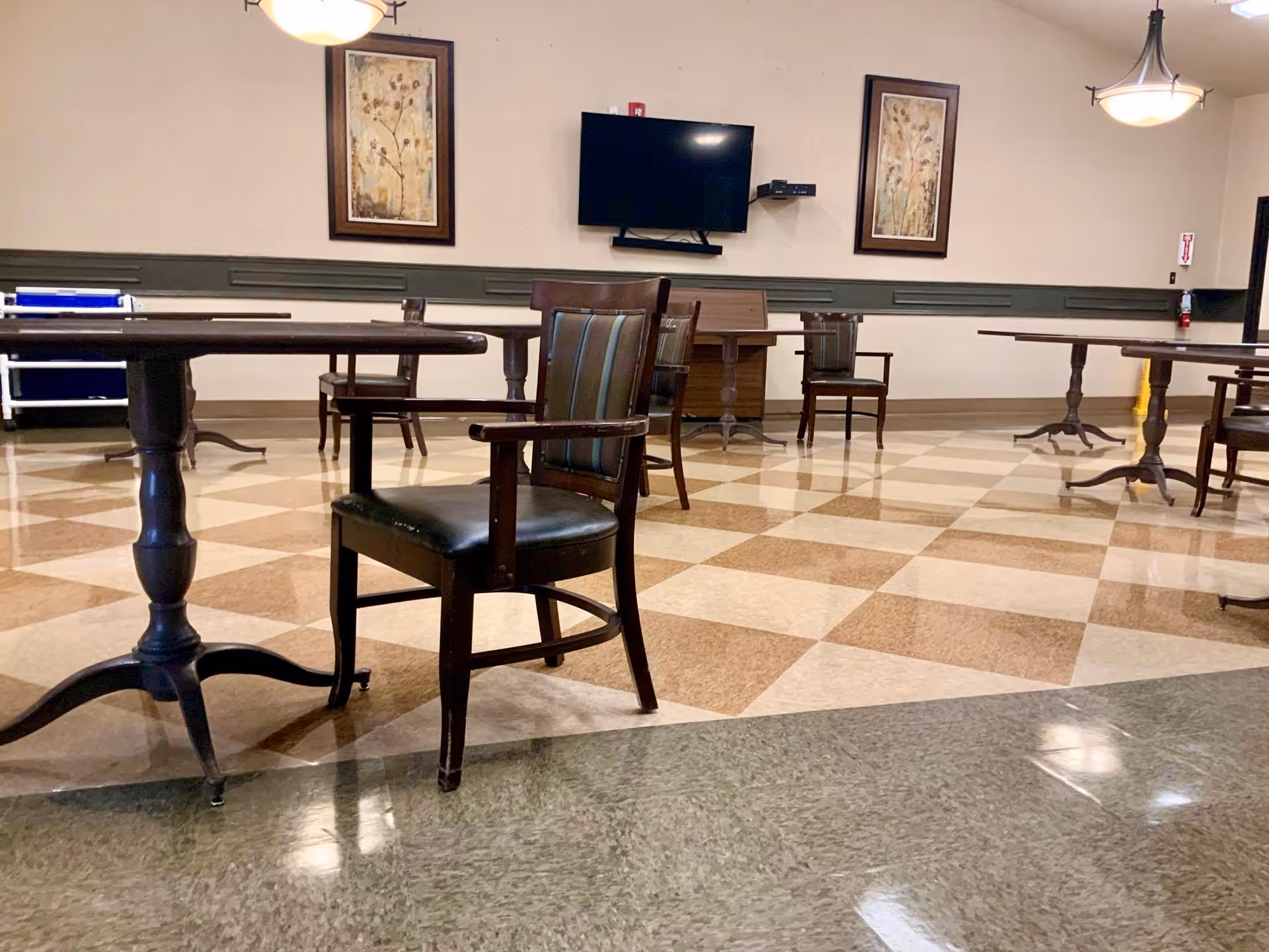 An empty common area with several wooden tables and chairs arranged on a checkered tile floor. The walls are beige with a dark green wainscoting, and there are two framed floral paintings hanging on the wall. A flat-screen TV is mounted on the wall between the paintings. Ceiling lights hang from above, and a fire extinguisher is visible near the door on the right.