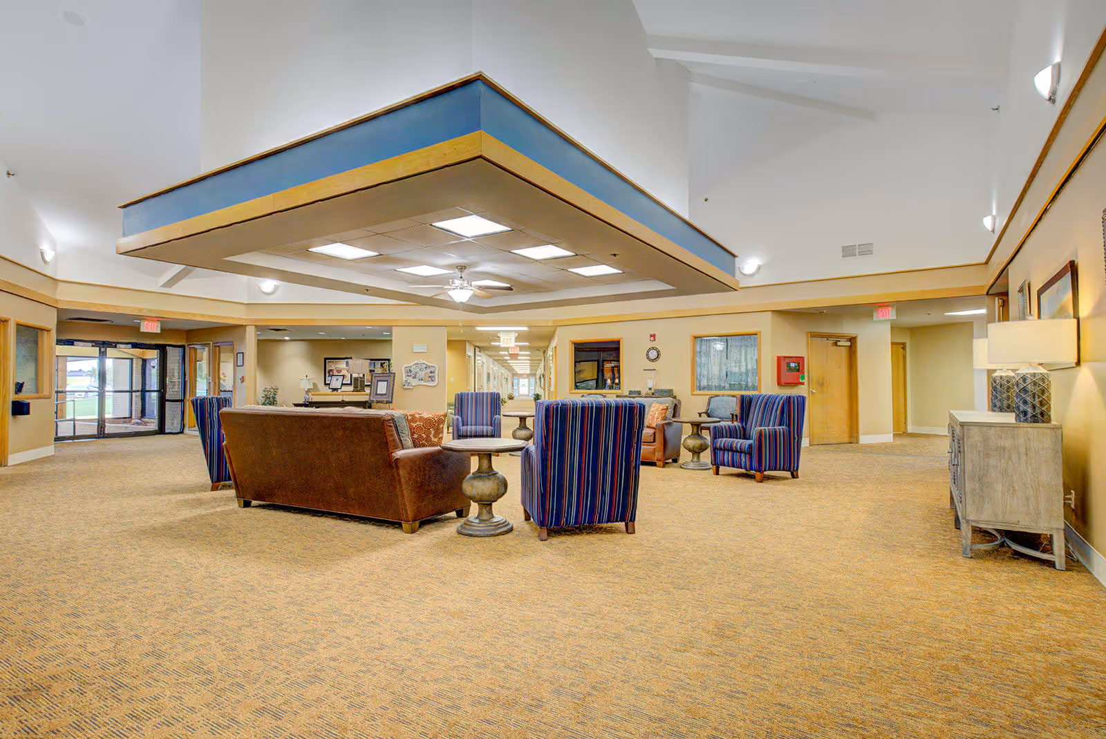 A spacious assisted living facility common area with a high ceiling and a central recessed ceiling section with lights and a ceiling fan. The room features a seating arrangement including a brown leather sofa, several striped armchairs, and round side tables. The walls are painted beige with wooden trim, and there are lamps and framed artwork on the walls. Doors and windows are visible along the walls, and exit signs are above the doors.