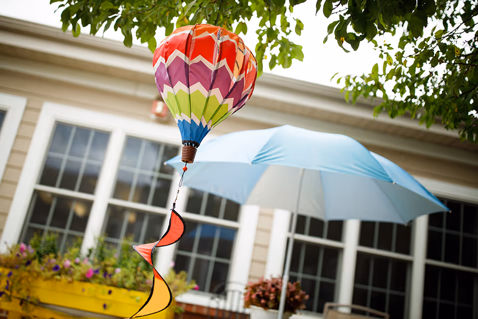 A colorful hot-air-balloon wind spinner hangs in front of a blue patio umbrella, windowed building, and planters.