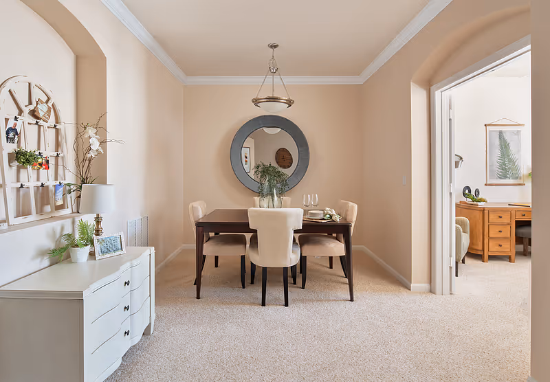 A dining room with a dark wooden table and four beige upholstered chairs. A round mirror hangs on the wall above the table, which has a vase with greenery and two wine glasses. To the left, there is a white dresser with a lamp, plants, and framed photos. An archway leads to another room with a wooden desk and chair, decorated with a framed leaf print on the wall.