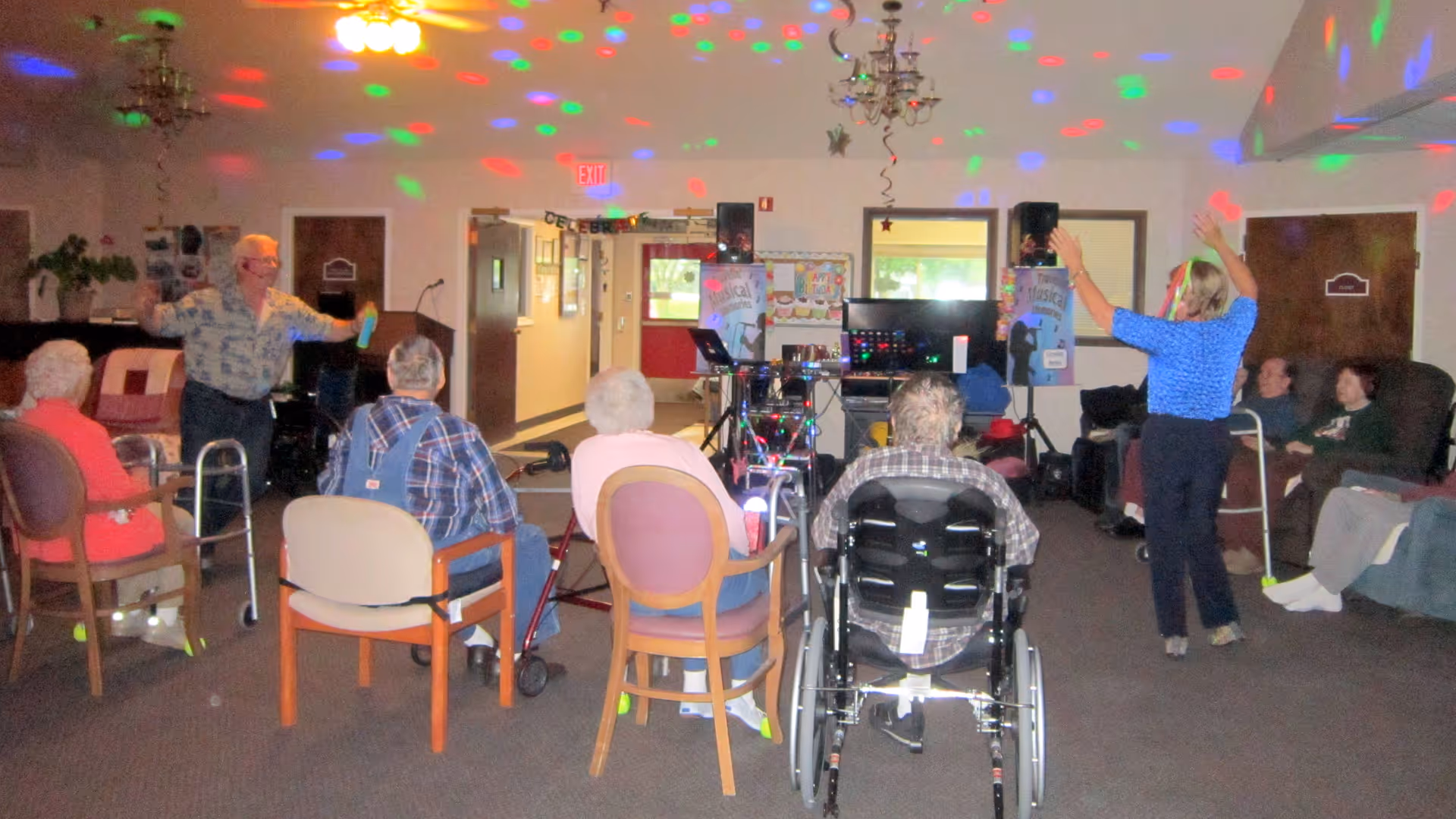 A group of elderly people seated in a common room with some in wheelchairs and others in chairs, participating in a group activity with two instructors standing and raising their arms. Colorful disco lights are projected on the ceiling and walls, creating a festive atmosphere.