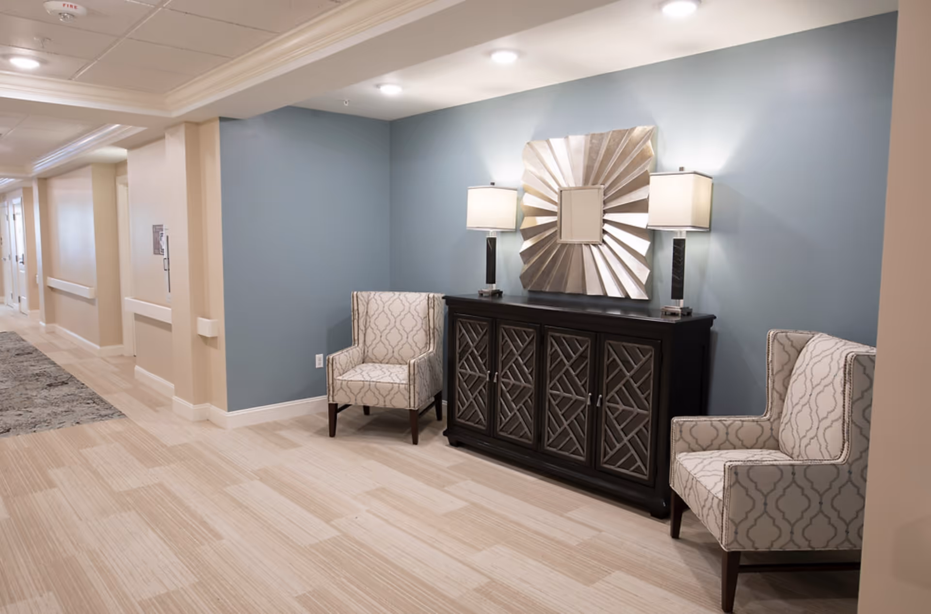 Hallway seating area with two patterned armchairs flanking a dark console table topped by lamps and a decorative mirror.
