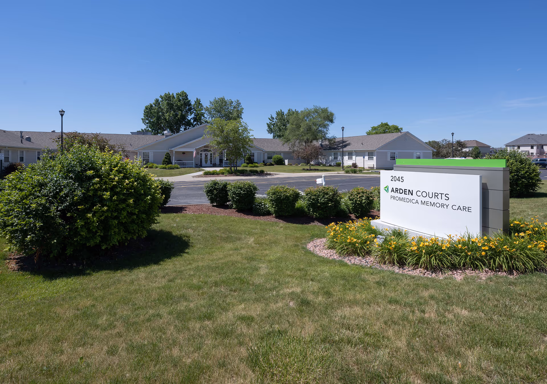 Front exterior of Arden Courts ProMedica Memory Care with landscaped lawn, entrance driveway, and a large sign in the foreground under a clear blue sky.