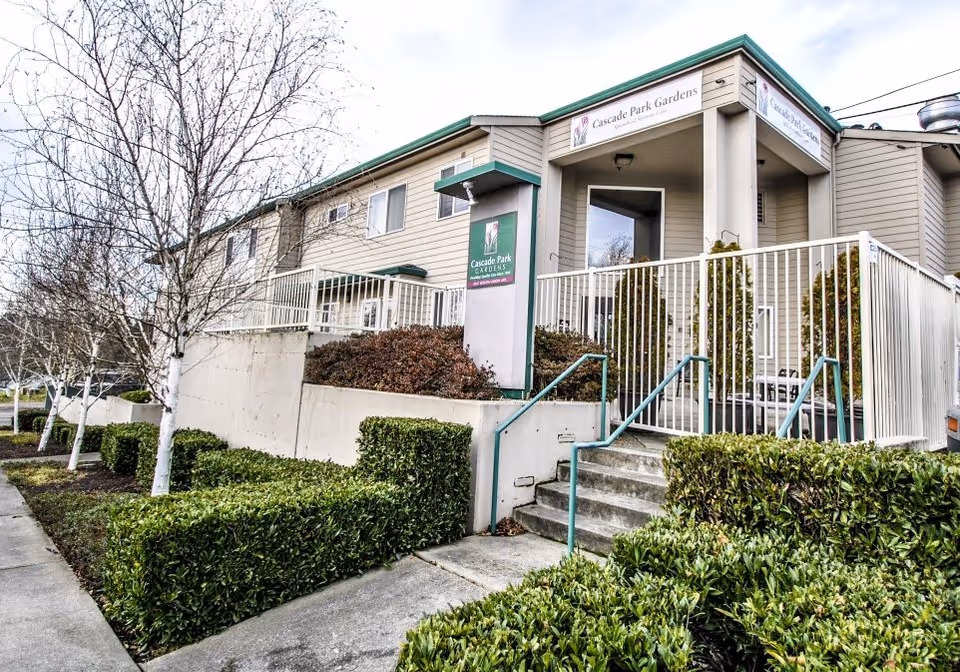 Exterior view of Cascade Park Gardens facility showing a two-story building with beige siding, green roof trim, and a fenced entrance with stairs leading up. There are trimmed bushes and leafless trees along the sidewalk in front of the building.