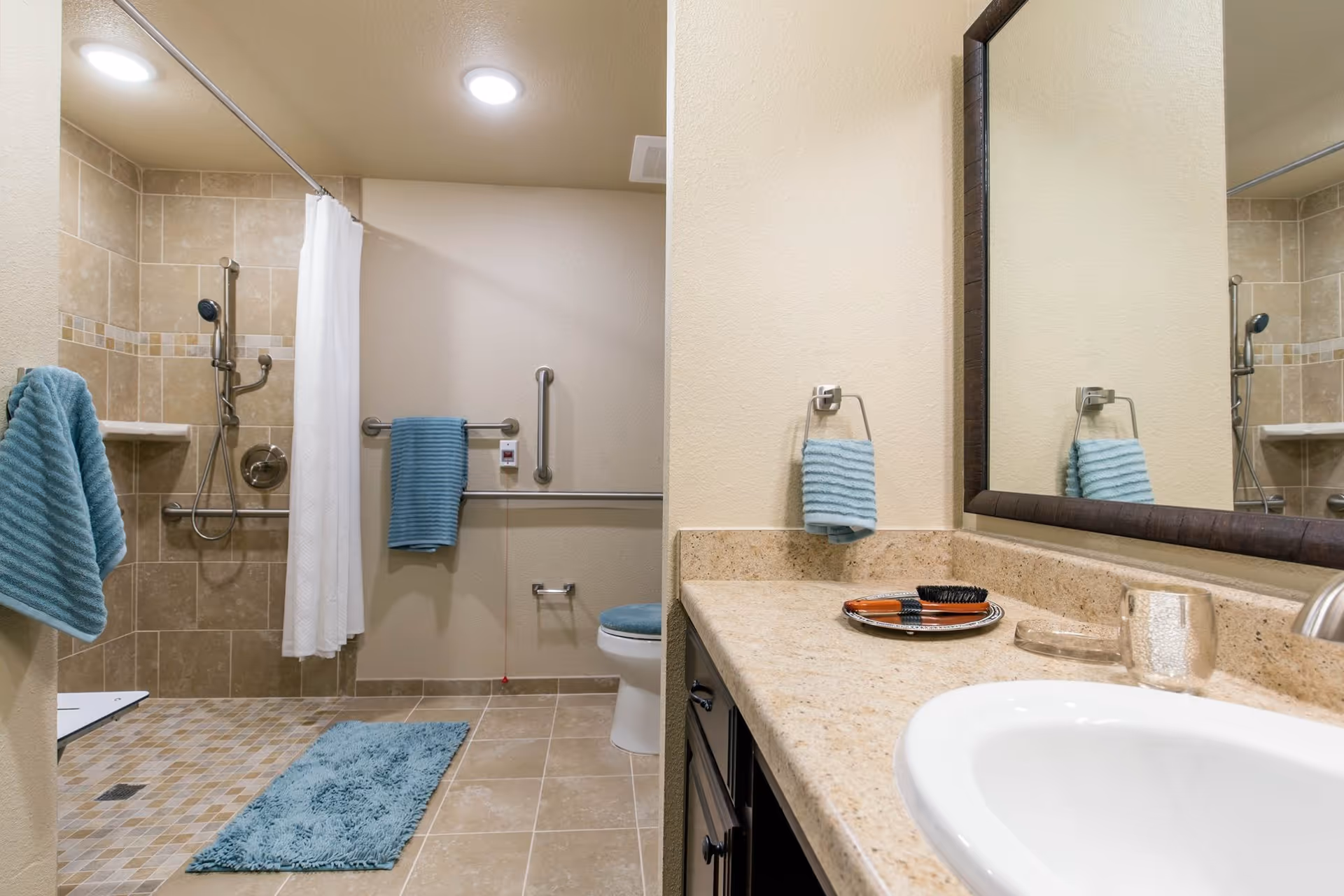 A clean and accessible bathroom with beige tiled floors and walls. The shower area has a white curtain, a handheld showerhead, and grab bars. There are blue towels hanging on the wall and a matching blue bath mat on the floor. A toilet with a blue seat cover is visible next to the shower. The countertop has a sink, a mirror, and a few small items including a hairbrush and a glass.