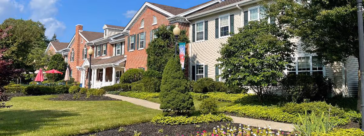 Front exterior of a senior living building with brick and siding facades, landscaped lawn, a walkway, and patio umbrellas.