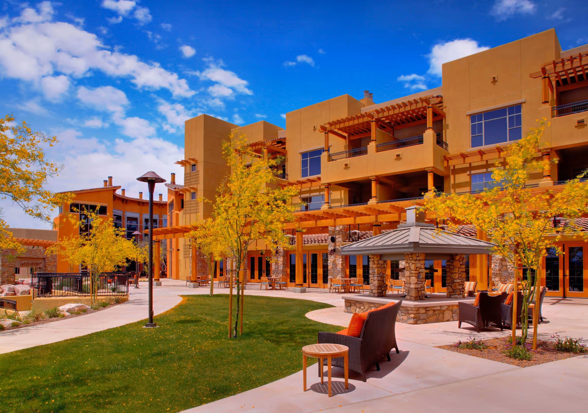 Courtyard seating area and landscaped grounds in front of a modern multi-story senior living building under a blue sky.