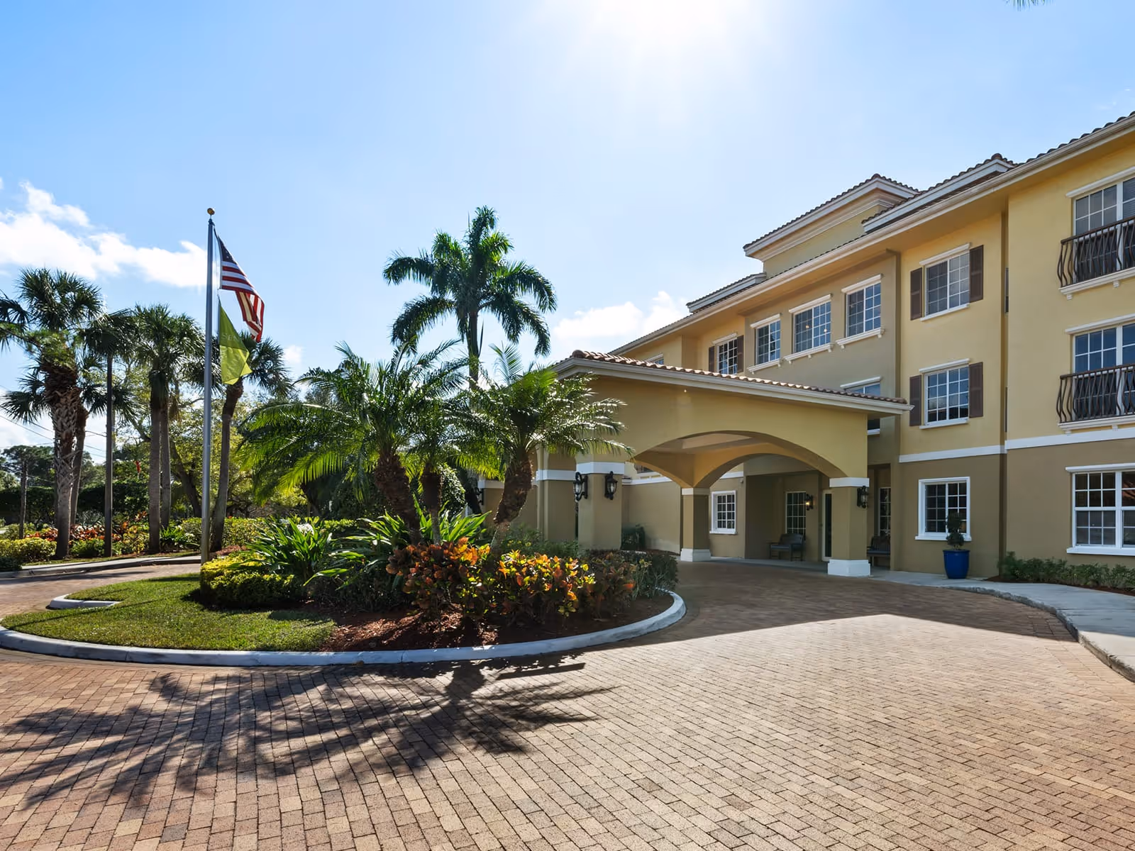Exterior view of a senior living facility with a covered entrance, palm trees, and landscaped greenery under a clear blue sky. Two flags are flying on flagpoles near the entrance.