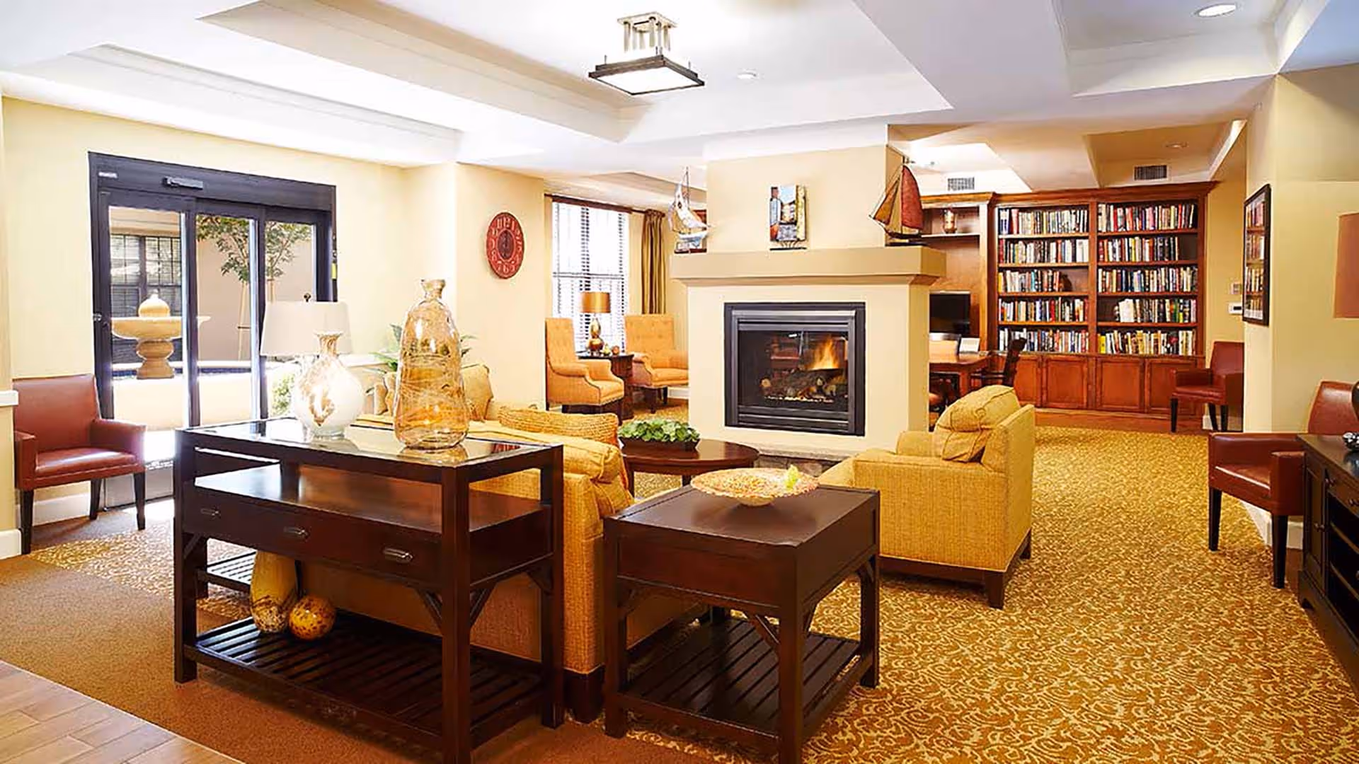 Well-lit communal lounge with sofas, armchairs, a central fireplace, bookshelves and side tables.