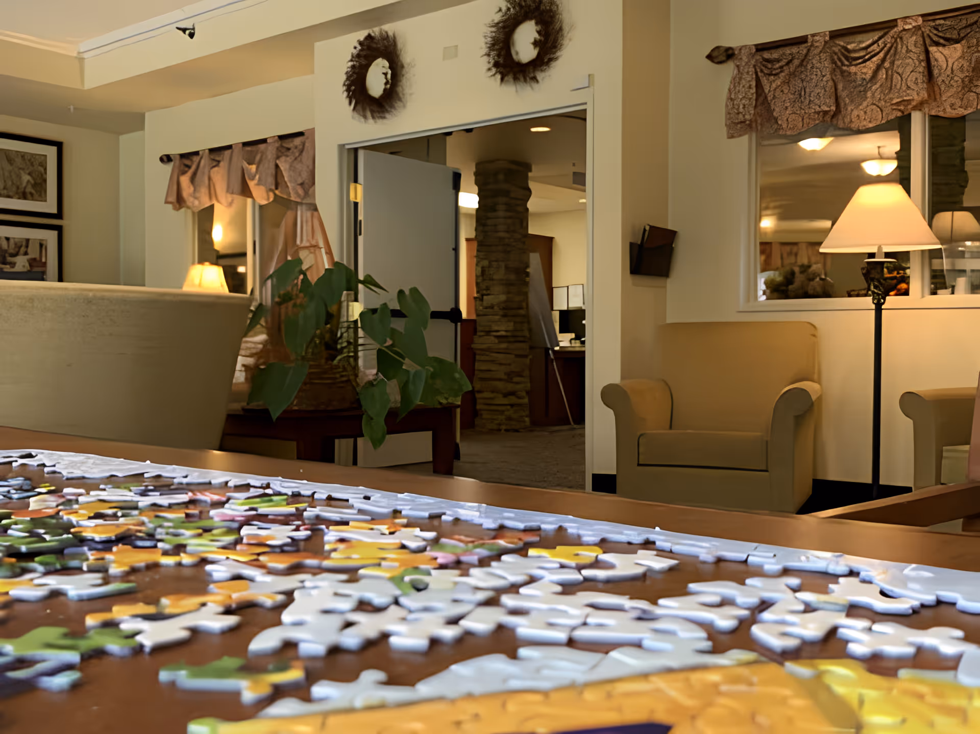 A cozy common area in a senior living facility with a table covered in scattered puzzle pieces in the foreground. The room features beige armchairs, a floor lamp, a potted plant, wall decorations, and a window with draped curtains. An open door reveals an adjacent room with a stone pillar and office equipment.