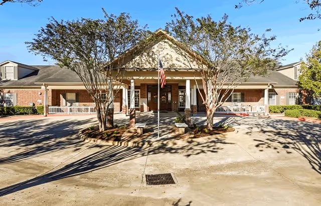 Front exterior view of a single-story brick building with a covered entrance supported by white columns. There are two trees in front of the entrance and an American flag on a flagpole in the center. The building has multiple windows and a paved driveway in front.