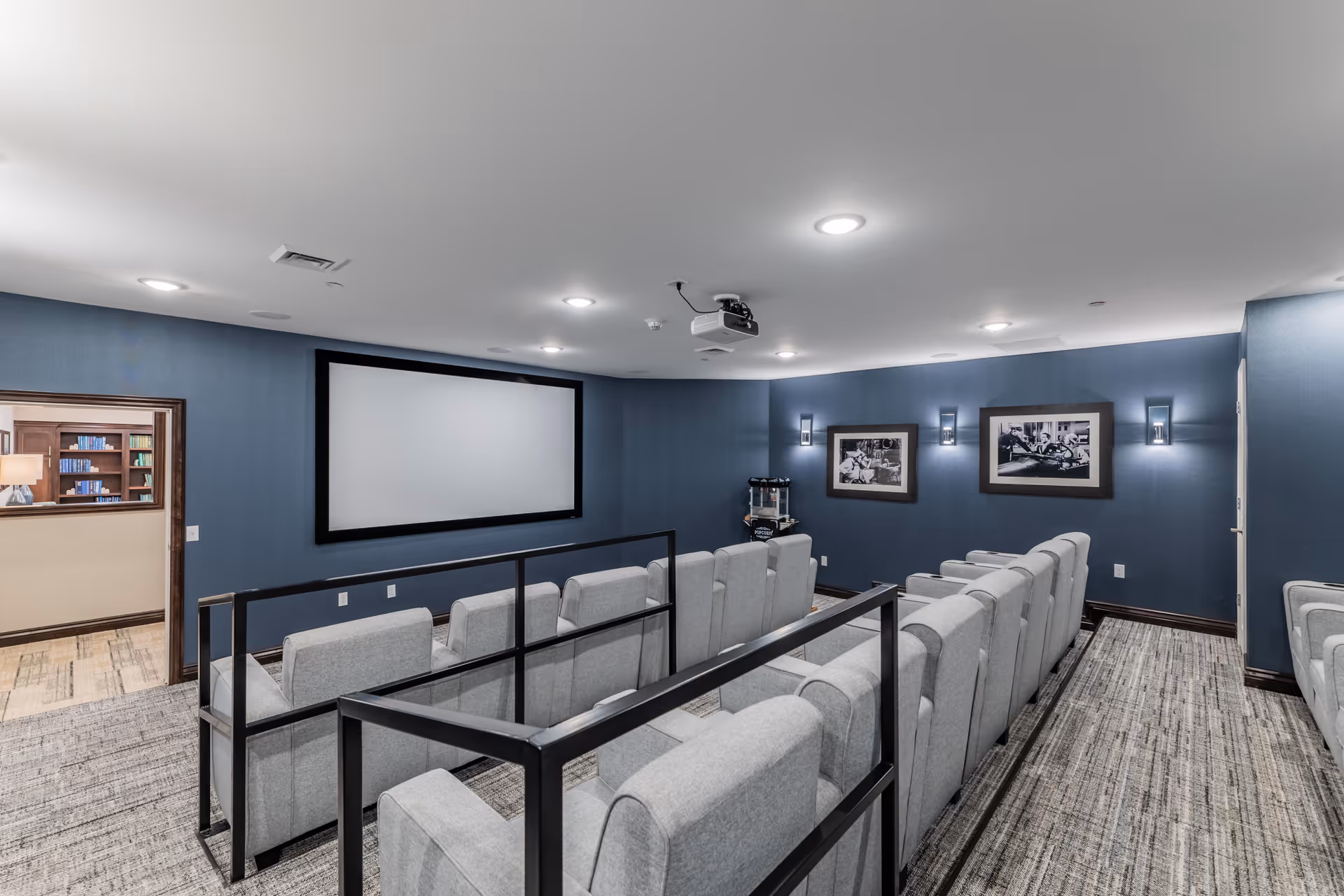 A small theater room with rows of gray upholstered seats facing a large blank projection screen on a blue wall. The room has a ceiling-mounted projector, wall sconces, and framed black and white photographs on the walls. There is a popcorn machine in the corner and an open doorway leading to a room with bookshelves.