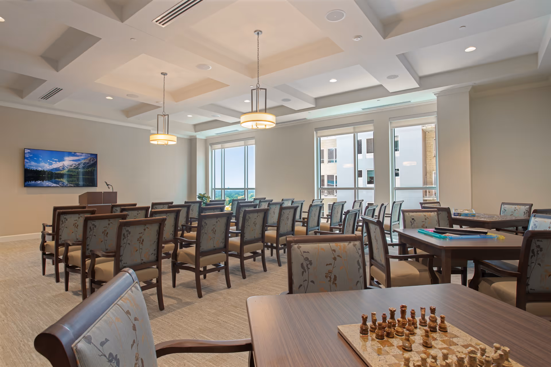 A well-lit meeting or activity room with multiple rows of chairs facing a podium and a wall-mounted TV screen displaying a scenic mountain and lake image. The room has large windows letting in natural light, several tables with chairs around them, and a chessboard set up on the nearest table.