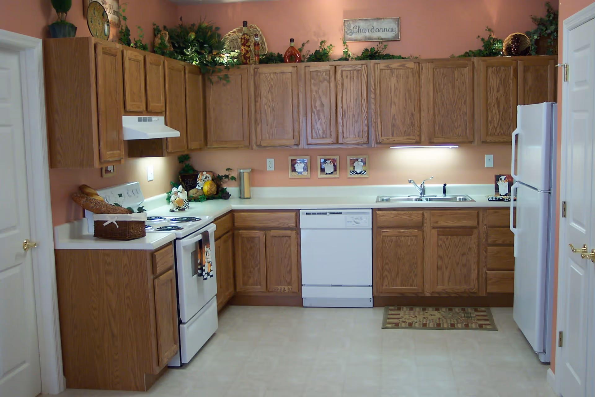 A kitchen with wooden cabinets, white countertops, a white stove with oven, a white dishwasher, and a white refrigerator. The walls are painted peach, and there are decorative plants and items on top of the cabinets. A small rug is placed in front of the sink area.