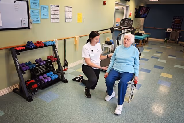 A senior woman seated on a chair holding small red dumbbells in each hand while a female caregiver or physical therapist kneels beside her, assisting with exercise in a fitness room equipped with various dumbbells, exercise equipment, and instructional posters on the wall.