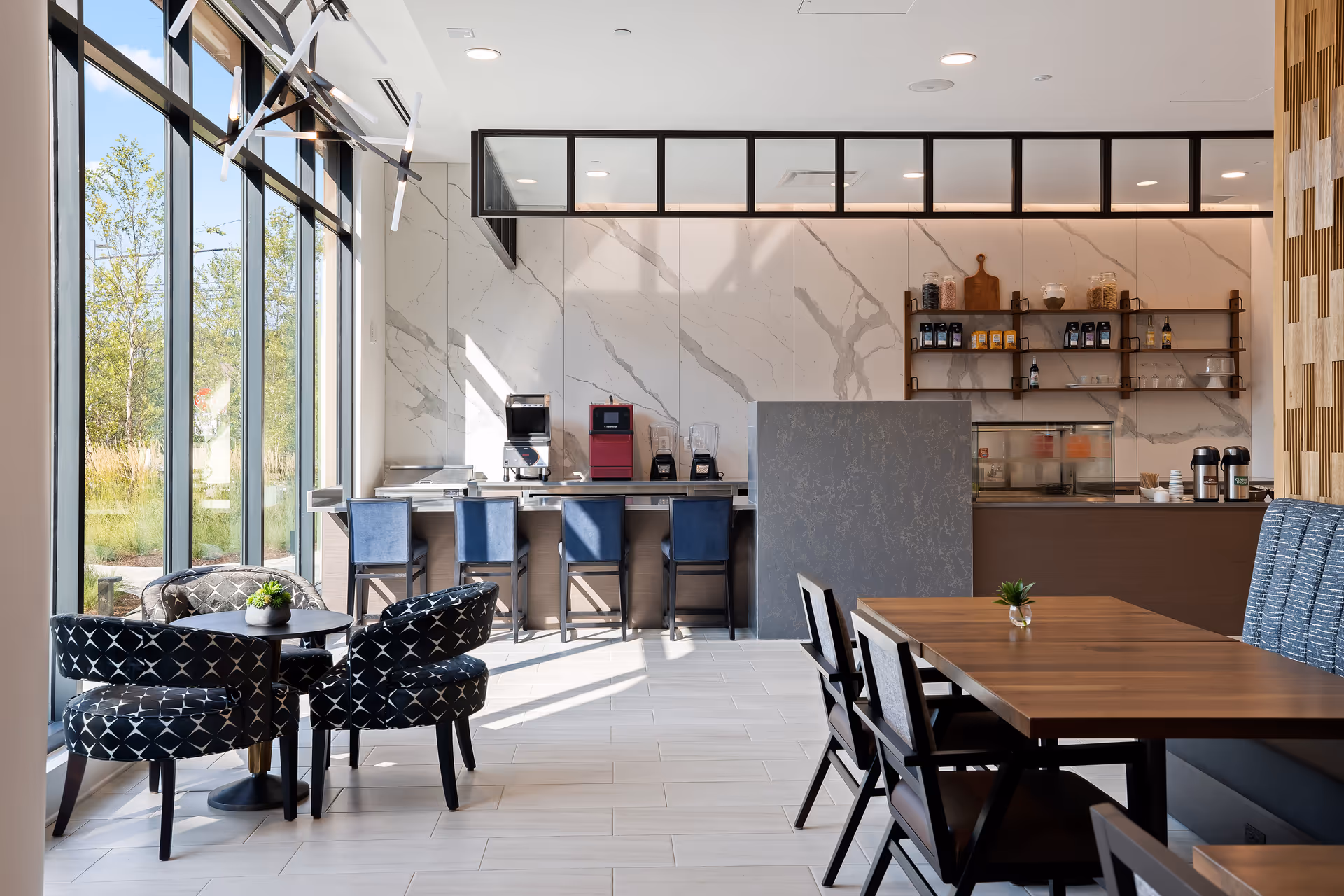 A modern dining area with large floor-to-ceiling windows letting in natural light. The space features a small round table with four patterned black chairs, a wooden rectangular table with chairs and bench seating, and a counter area with stools. The back wall has marble-like panels, shelves with various containers, and coffee and blender machines.