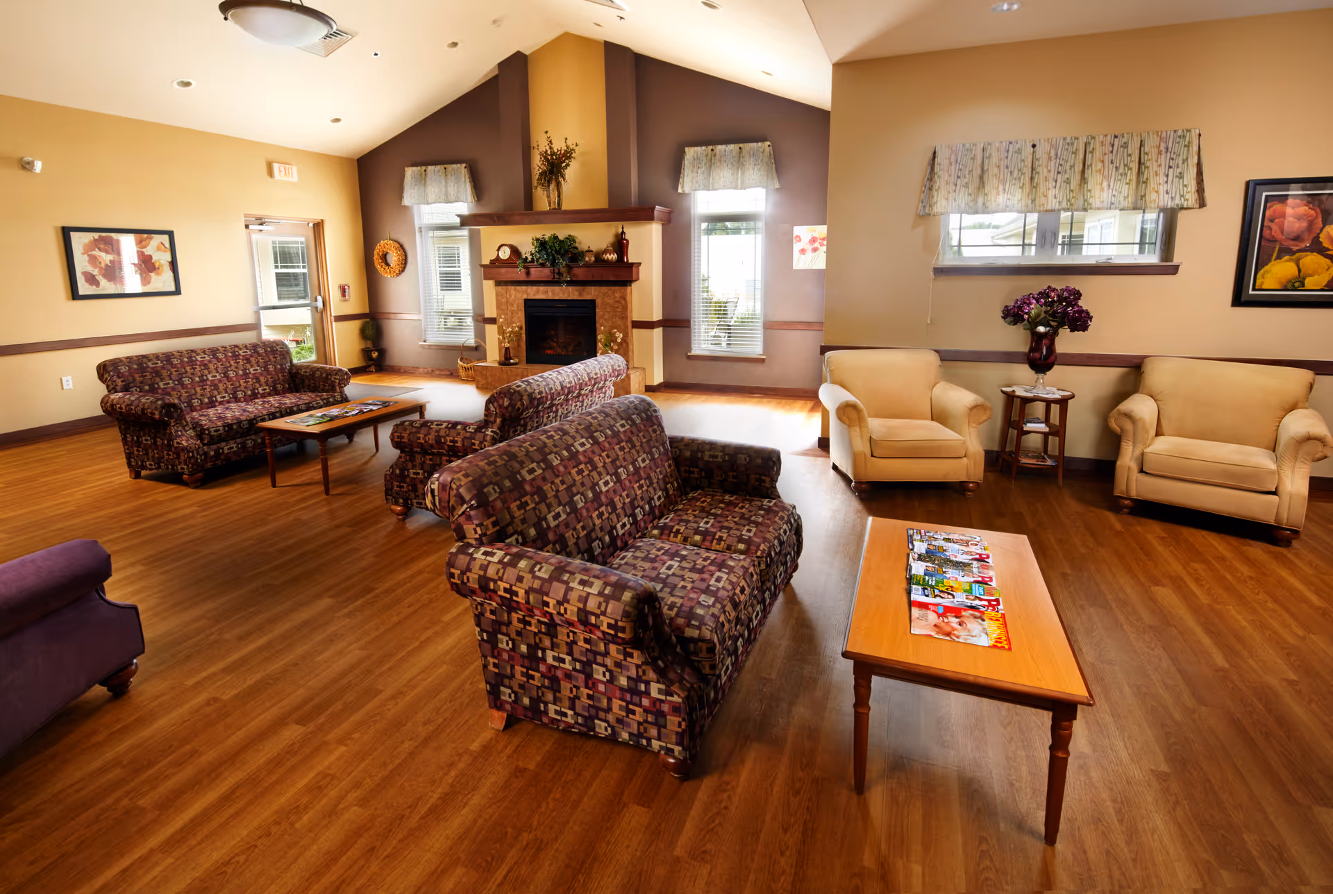 A spacious living room area with wooden flooring, featuring multiple upholstered sofas and armchairs arranged around a wooden coffee table with magazines on it. The room has a fireplace with decorative plants and vases on the mantle, windows with valances, framed artwork on the walls, and soft lighting from ceiling fixtures.
