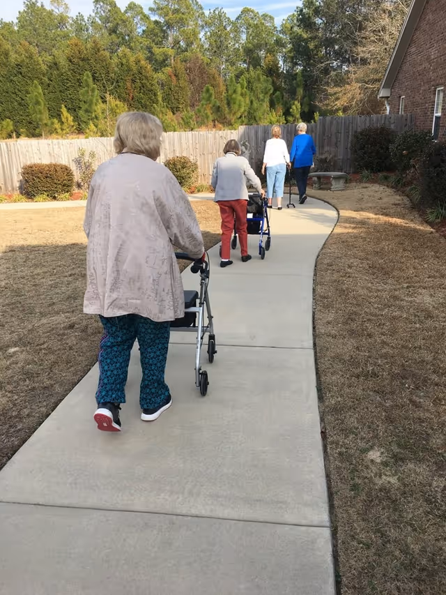 Four elderly women walking on a curved concrete pathway outdoors, each using a walker for support. The path is bordered by grass and bushes, with a wooden fence and trees in the background. The women are dressed in casual clothing and are walking away from the camera.