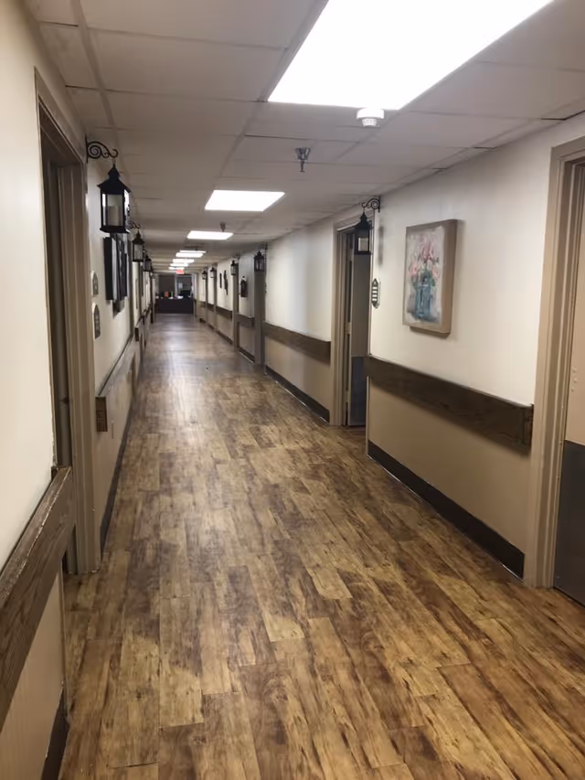 Long, well-lit interior hallway with wood-look flooring, handrails and doors lining both sides.