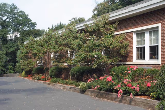 Exterior view of a brick building with white-framed windows, surrounded by a landscaped garden featuring small trees, bushes, and pink flowers along a paved driveway.