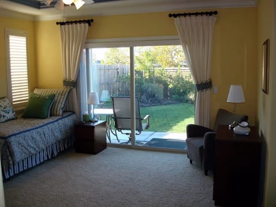 A cozy room with yellow walls featuring a daybed with multiple pillows on the left, a small wooden nightstand with a lamp beside it, and a sliding glass door leading to a green backyard with patio furniture. On the right side, there is a dark armchair next to a wooden dresser with a lamp and decorative items on top. The room has beige carpet and white curtains framing the sliding door.