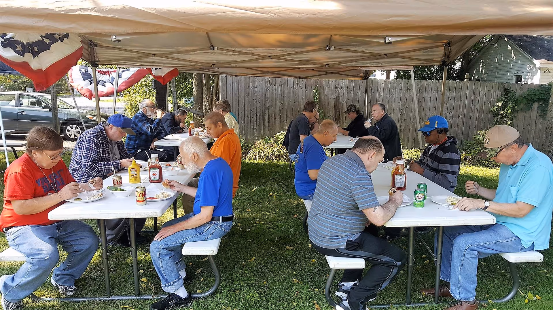 A group of elderly adults sitting at two white picnic tables under a canopy tent outdoors, eating a meal together. The tables have condiments like ketchup and mustard, and some soda cans. The setting is a grassy area with a wooden fence and some trees in the background.