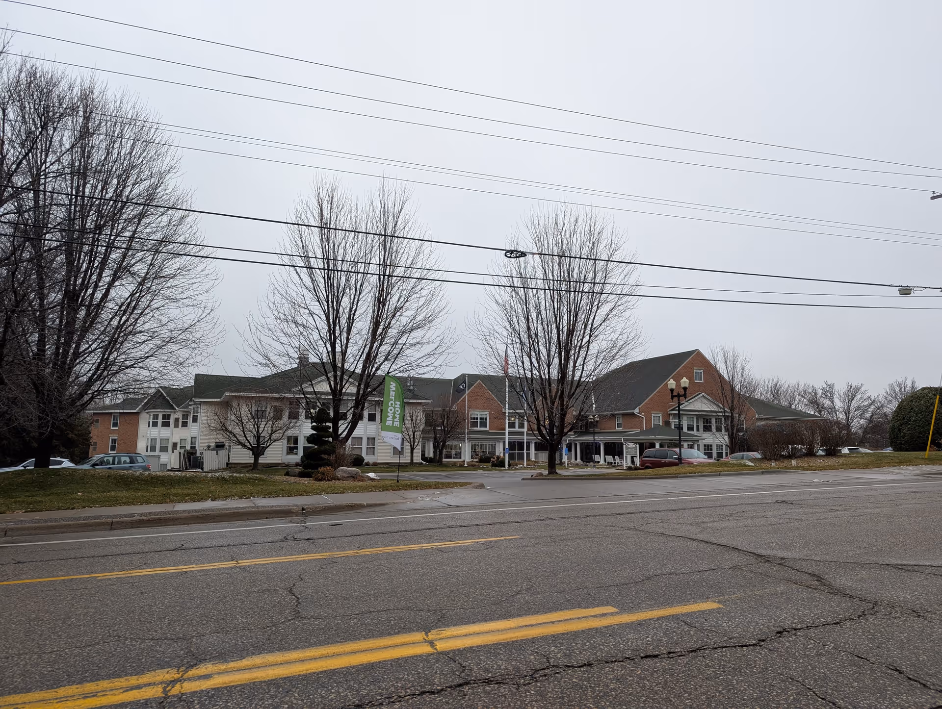 Exterior view of Heathers Manor, a senior living facility, showing a large building with a mix of brick and white siding. There are leafless trees in front, a green flag with 'HOME MET COWE' text, several parked cars, and a cloudy sky overhead.