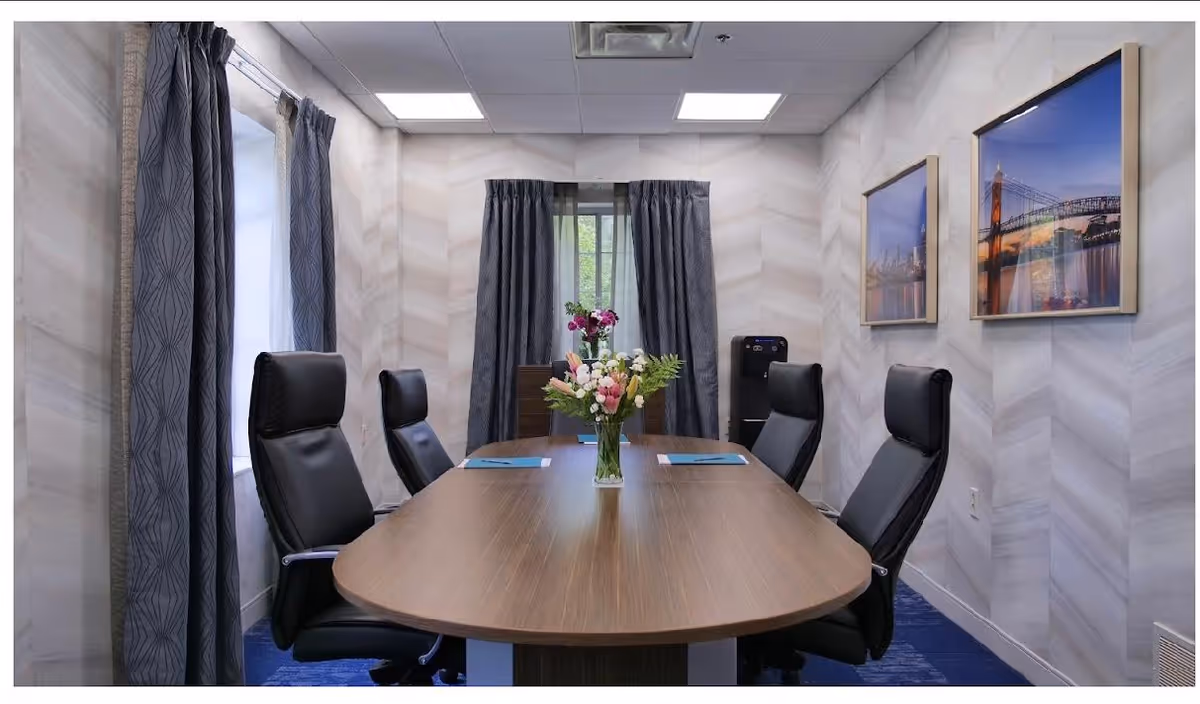 A small conference room with a wooden oval table surrounded by six black leather office chairs. There are two windows with dark curtains, a vase with flowers on the table, two framed pictures of cityscapes on the wall, and a water dispenser in the corner.
