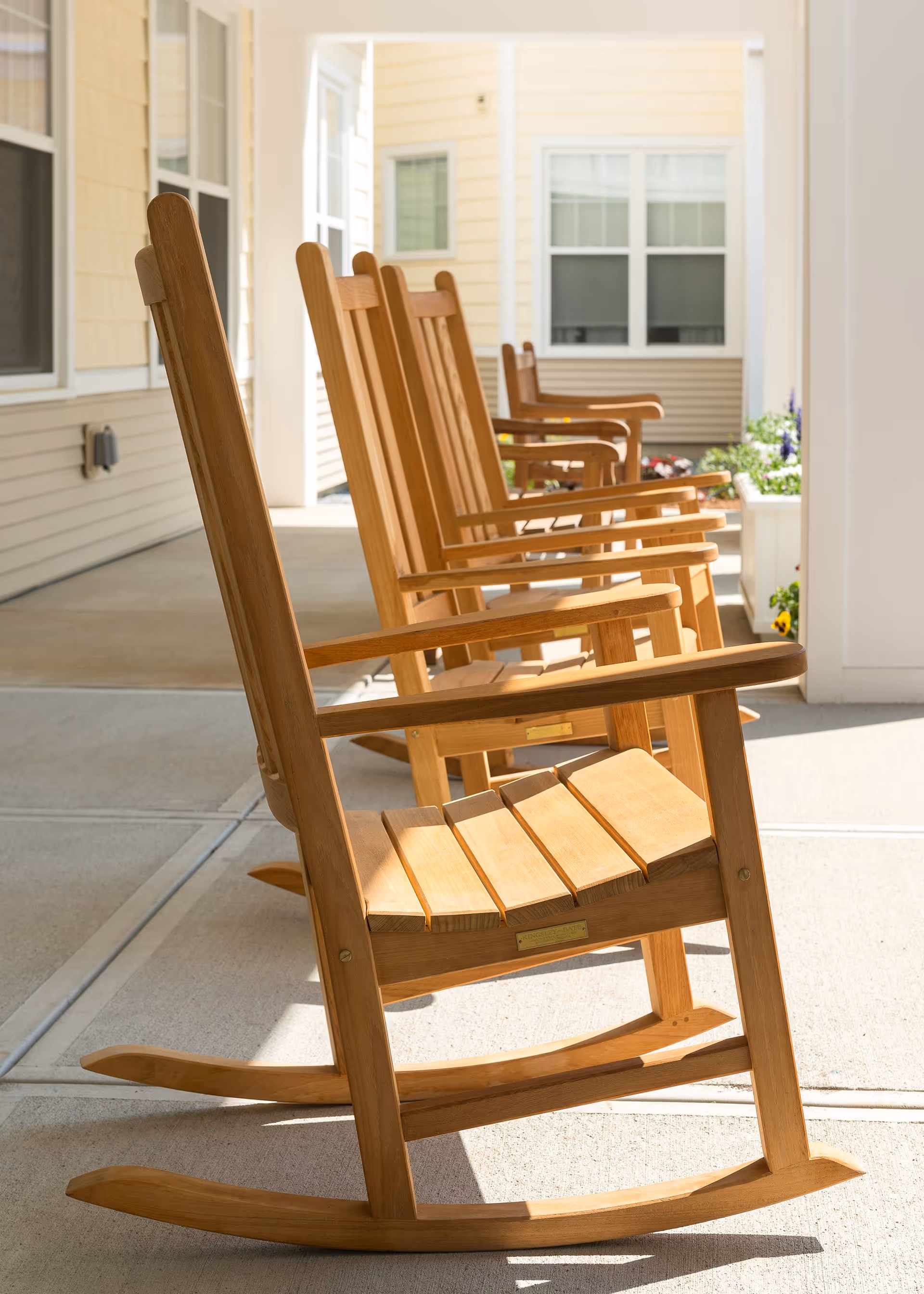 A row of wooden rocking chairs lined up on a covered porch with beige siding and windows in the background, with sunlight casting shadows on the concrete floor.