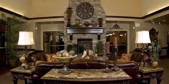 A cozy living room area with two brown leather sofas facing a stone fireplace. The fireplace has a large clock mounted above it and decorative items on the mantel. There are two table lamps on side tables flanking the sofas, and a coffee table with decorative objects in the center. The room has warm lighting and plants placed around the space.
