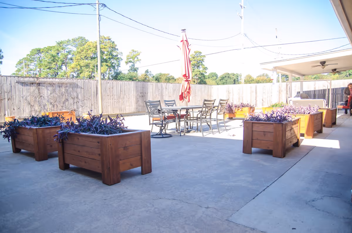 Outdoor patio area with several wooden planter boxes containing purple plants, a metal table with six chairs, and a closed red umbrella in the center. The area is enclosed by a wooden fence with trees visible beyond it. There is a covered section with ceiling fans on the right side and a person sitting in the background.