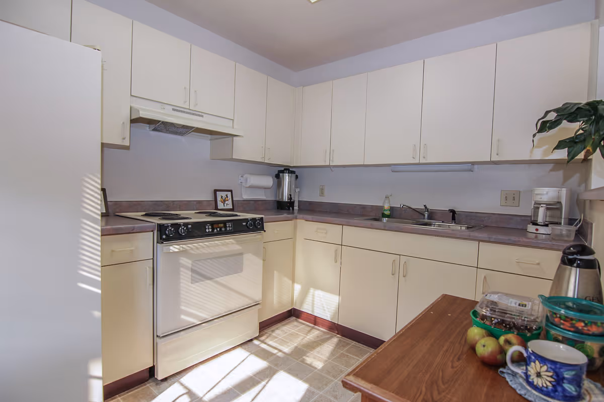 A bright kitchen with white cabinets and countertops. There is a white stove with an oven, a sink with a faucet, and various kitchen items including a coffee maker, a water dispenser, a thermos, a bowl of fruit, and a mug on a wooden table. Sunlight streams through a window, casting shadows on the floor and cabinets.