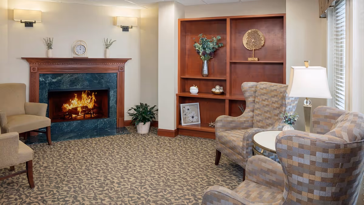 Cozy living room area with a lit fireplace surrounded by a green marble and wooden mantel. The room features patterned carpet, two beige armchairs with a checkered pattern, a small round table with a lamp and a small plant, a wooden built-in bookshelf with decorative items, and two wall-mounted lights above the fireplace.