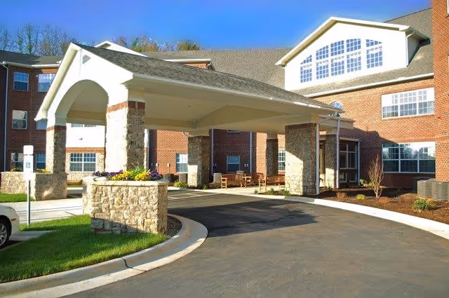 Covered drive-up porte-cochere entrance of a brick senior living building with benches and landscaped planters.