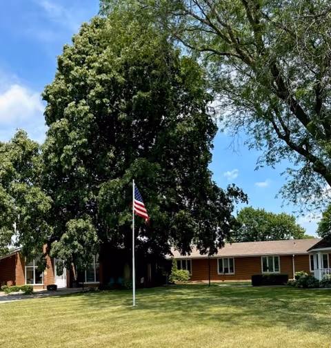 Single-story brick building front with a flagpole on a grassy lawn and large trees under a blue sky.