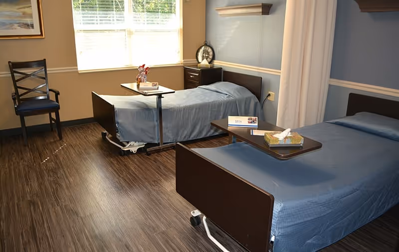 A room with two hospital-style beds covered with blue bedspreads, each with an overbed table. There is a wooden chair with armrests near a window with blinds, and a small nightstand with a clock and decorative items. The floor is wood-patterned, and the walls are painted in light colors with a chair rail molding.