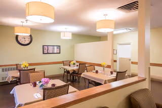 A dining area with several tables covered with beige tablecloths, each set with cups, napkins, and small flower arrangements. The room has light green walls, a large wall clock, framed artwork, and ceiling lights with round beige lampshades.