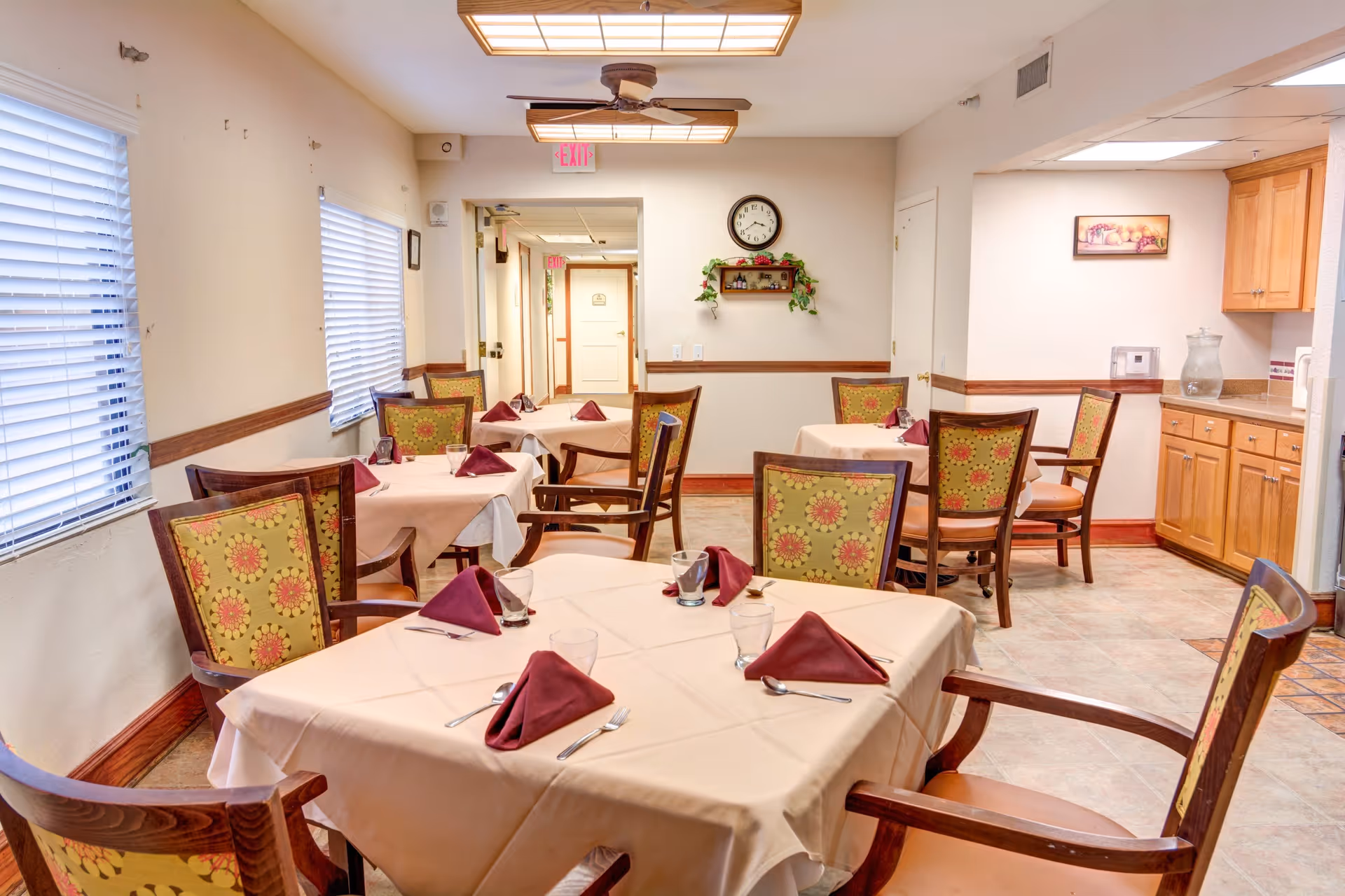Dining room with several tables covered with beige tablecloths, each set with folded maroon napkins, glasses, and silverware. The chairs have wooden frames with floral patterned upholstery. The room has windows with blinds on the left wall, a ceiling fan with lights, a clock on the far wall above a small shelf with decorative items, and a kitchenette area with wooden cabinets on the right.