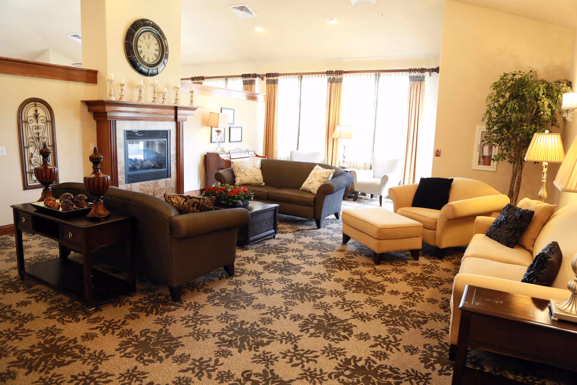 A spacious and well-lit living room area with multiple sofas and armchairs arranged around a central coffee table. The room features a patterned carpet, a fireplace with decorative candles on the mantle, a large clock above the fireplace, and large windows with curtains allowing natural light to fill the space. There are also several lamps and a large potted plant in the corner.
