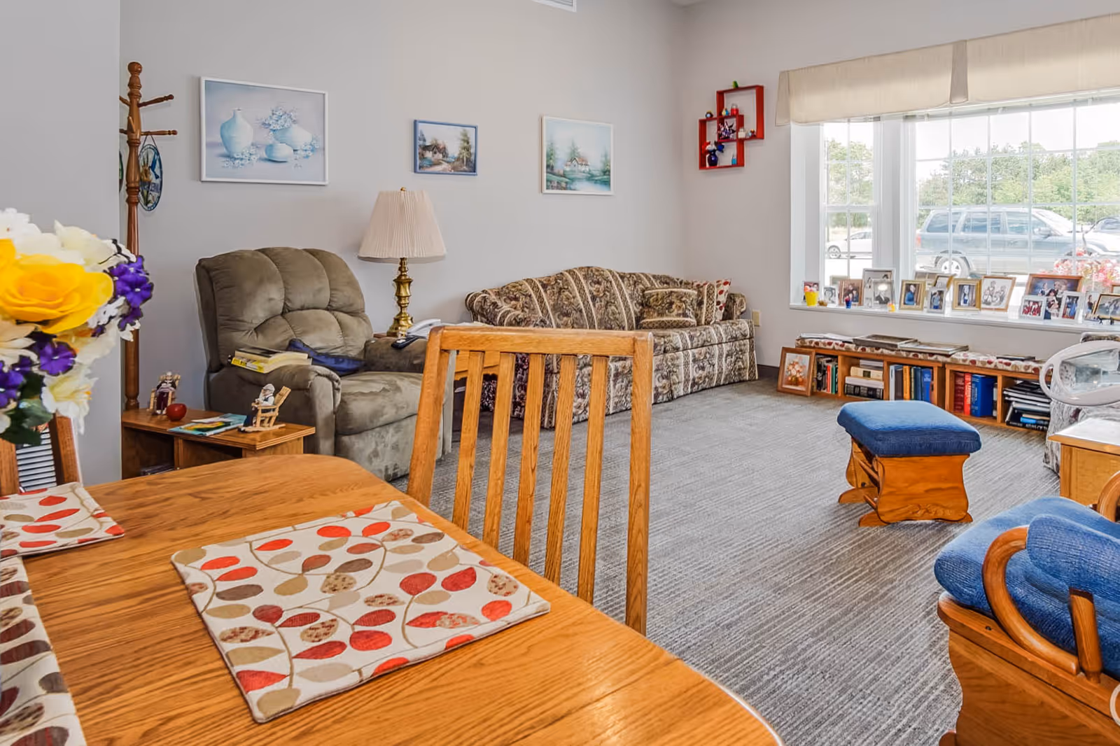 Bright assisted living common room with a dining table in the foreground, reclining chair, patterned sofa, ottoman, and a large window lined with framed photos.