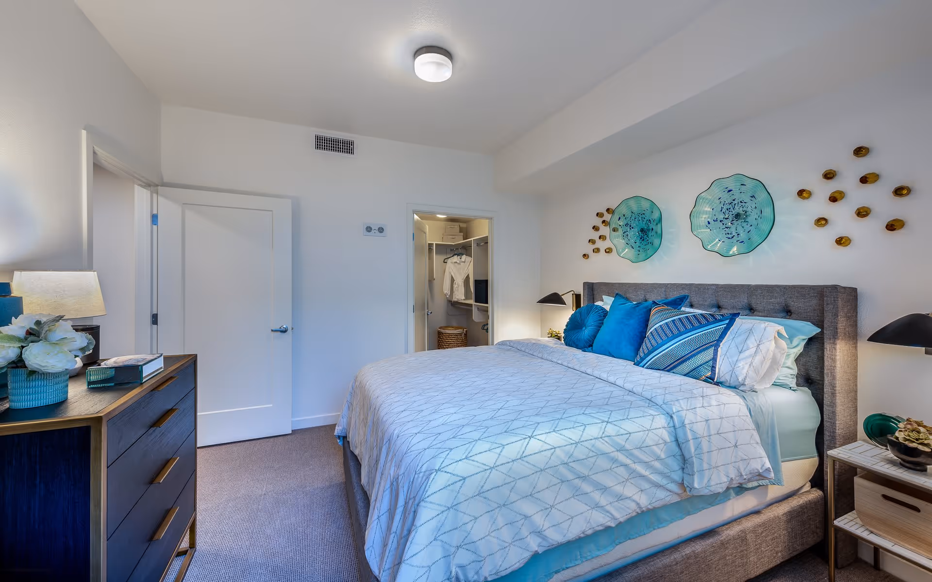 A modern bedroom with a large bed featuring blue and white bedding and multiple blue pillows. The room has a gray upholstered headboard, two black bedside lamps on either side, and decorative wall art above the bed consisting of two large blue glass plates and small gold accents. There is a wooden dresser with a lamp and flowers on the left side, and an open door leading to a closet with hanging clothes and storage baskets. The walls are white, and the room is well-lit with a ceiling light.